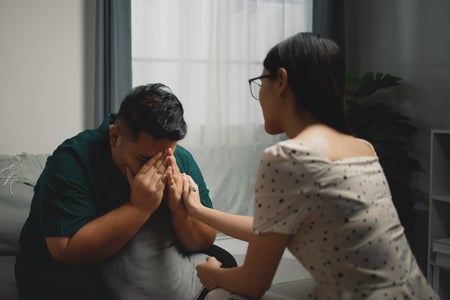 Woman places her hand on the arm and hand of a man as he sits on a couch with his hands covering his face in distress