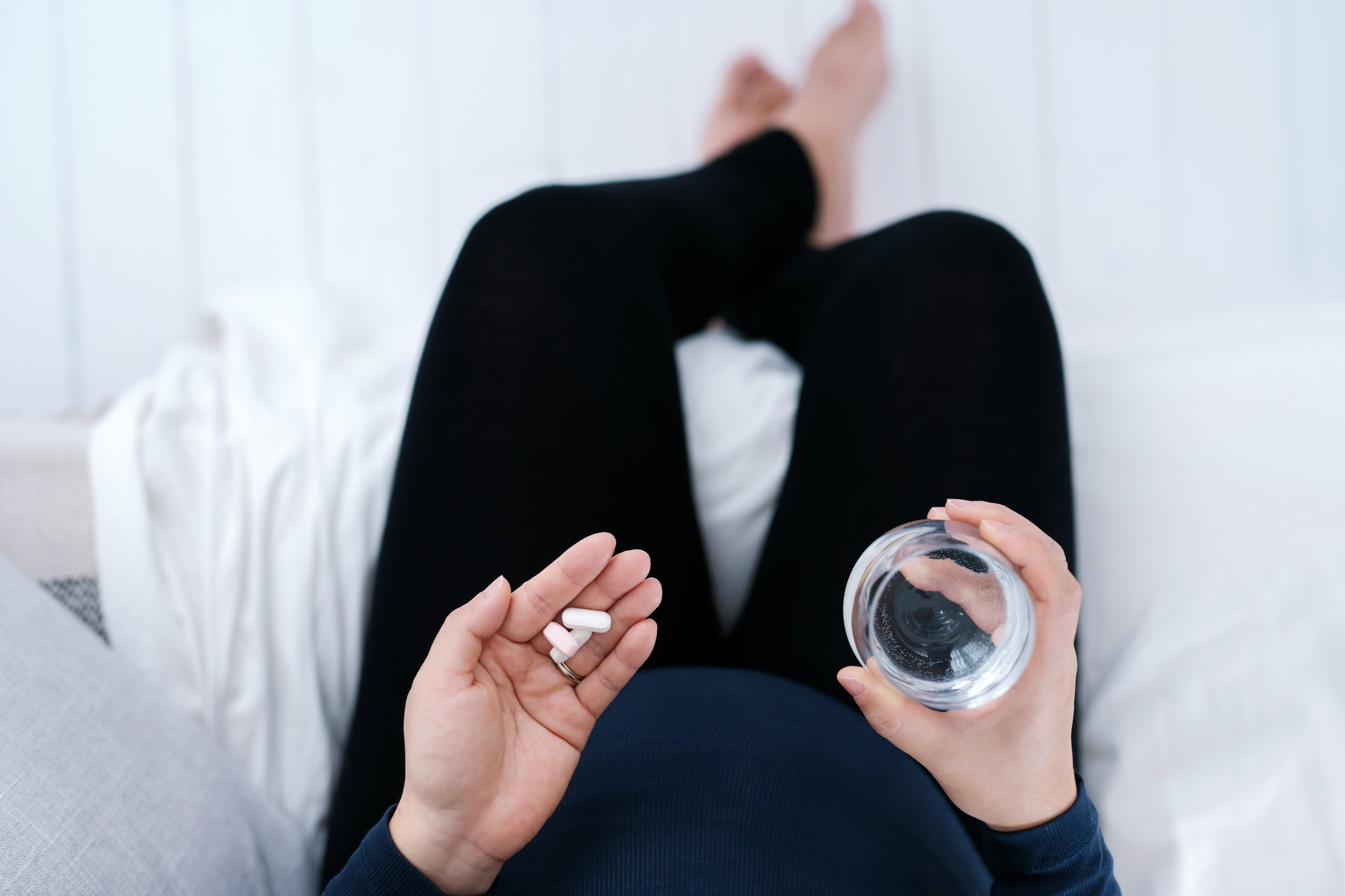 Pregnant woman's lower body in black pants on bed cross legges holding vitamins and a glass of water.