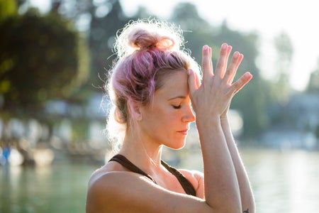 Woman with dyed hair meditating at a lake with open hands at forehead.