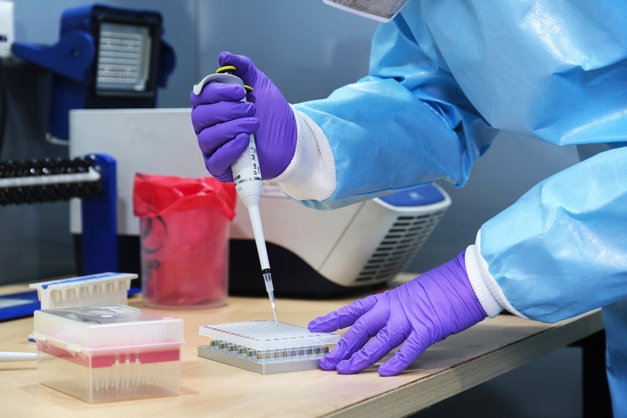 A healthcare worker wearing purple gloves and a blue full body protection suit uses a pipette to process Covid-19 test samples