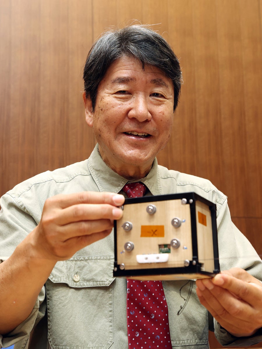 Asian astronaut with beige shirt and red tie in front of wood panel wall holding a small box wooden satellite.