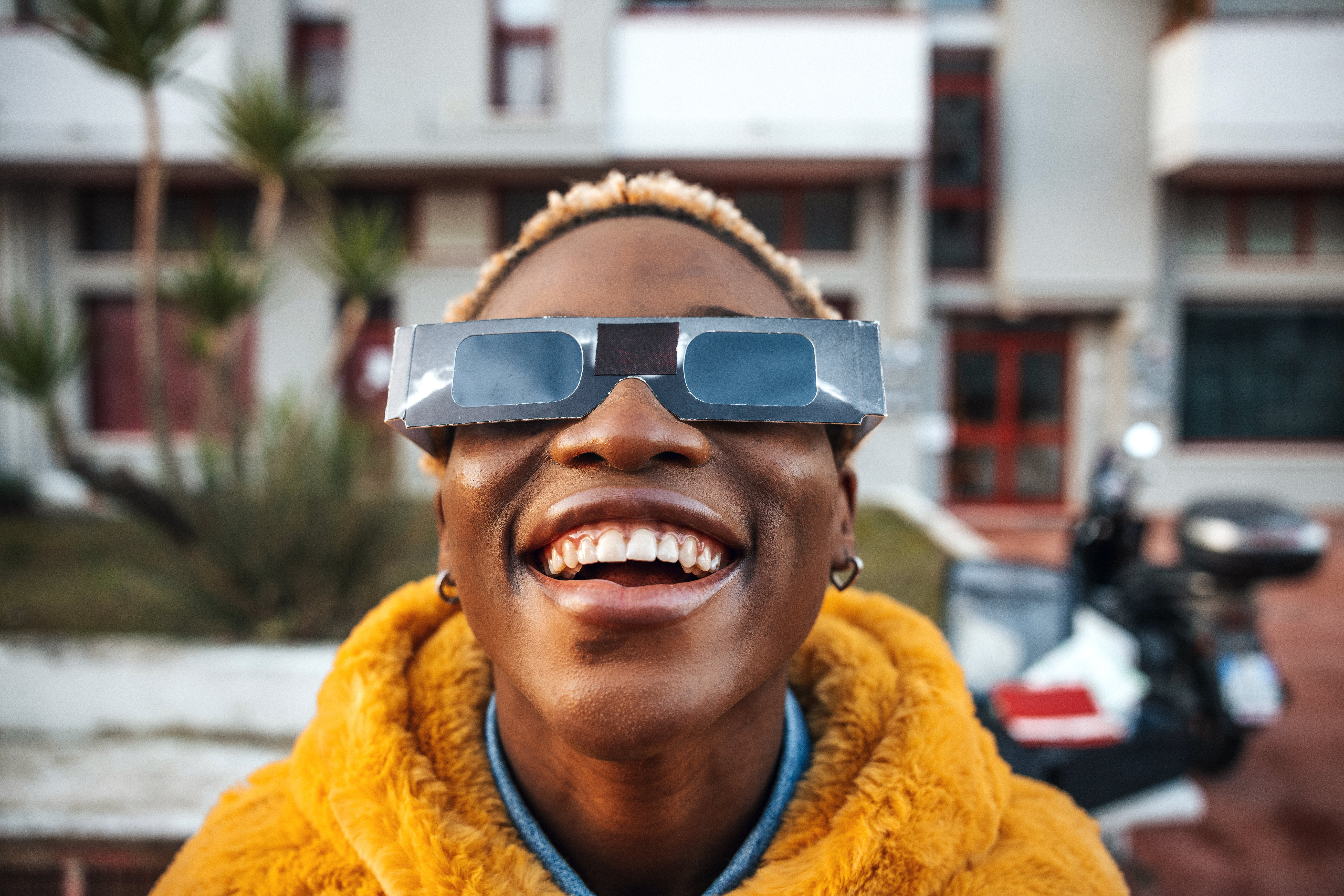 Young black woman wearing eclipse glasses and bright yellow jacket smiles while looking upward