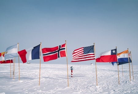 A series of national flags, including Argentina, France, Norway, the U.S., and Chile surround a small red and white striped pole with a round metal top on flat, white snow landscape against a light blue sky