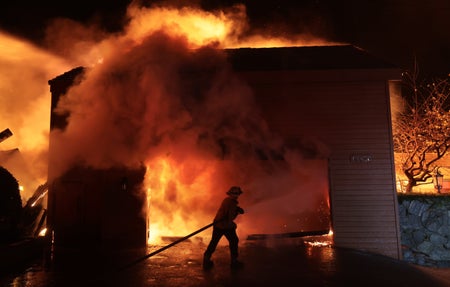 A firefighter battles a house fire surrounded by heavy smoke clouds within a building structure.