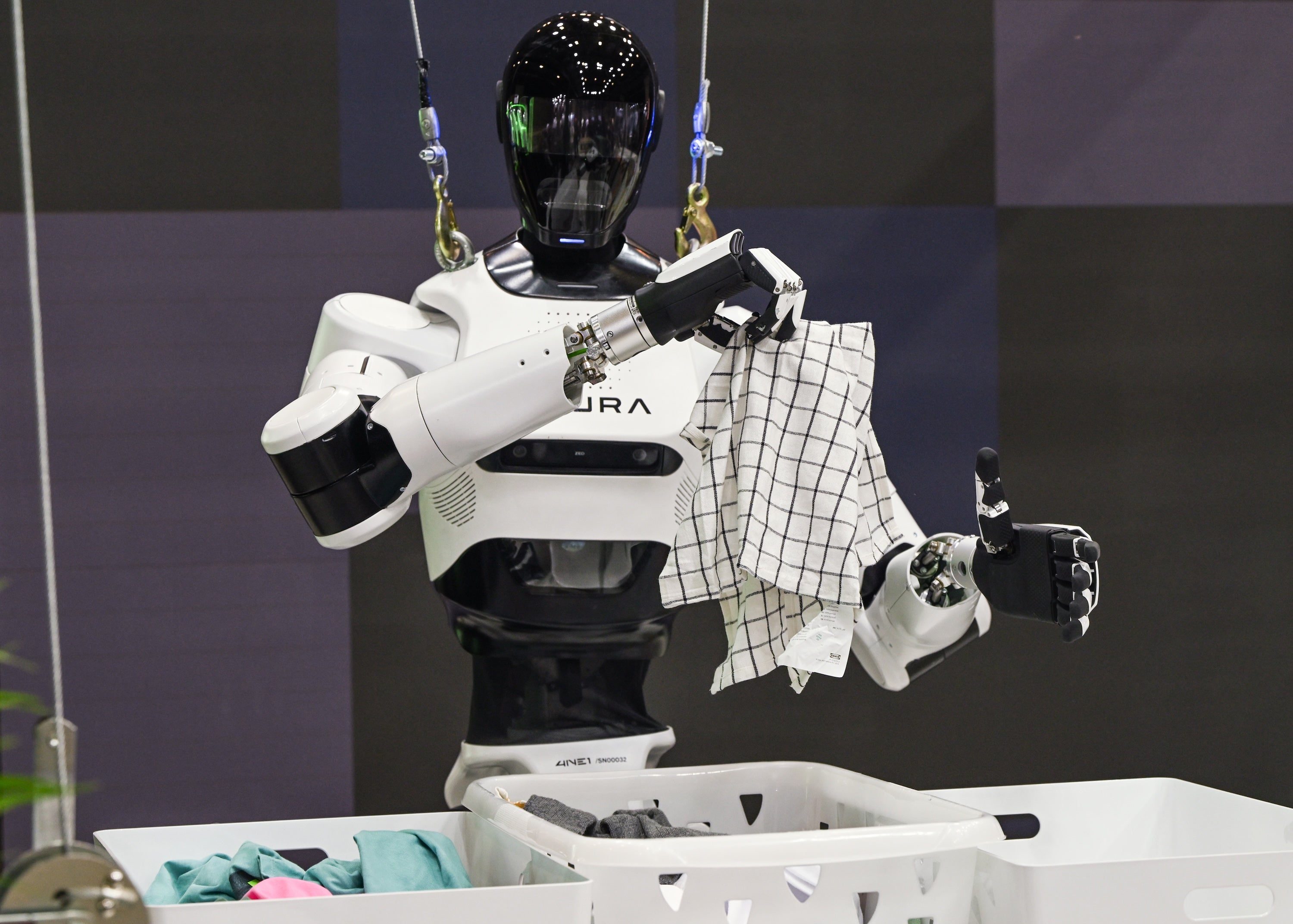 Humanoid robot with black visor folds laundry, holding checkered towel beside basket, suspended in lab with cables and equipment.