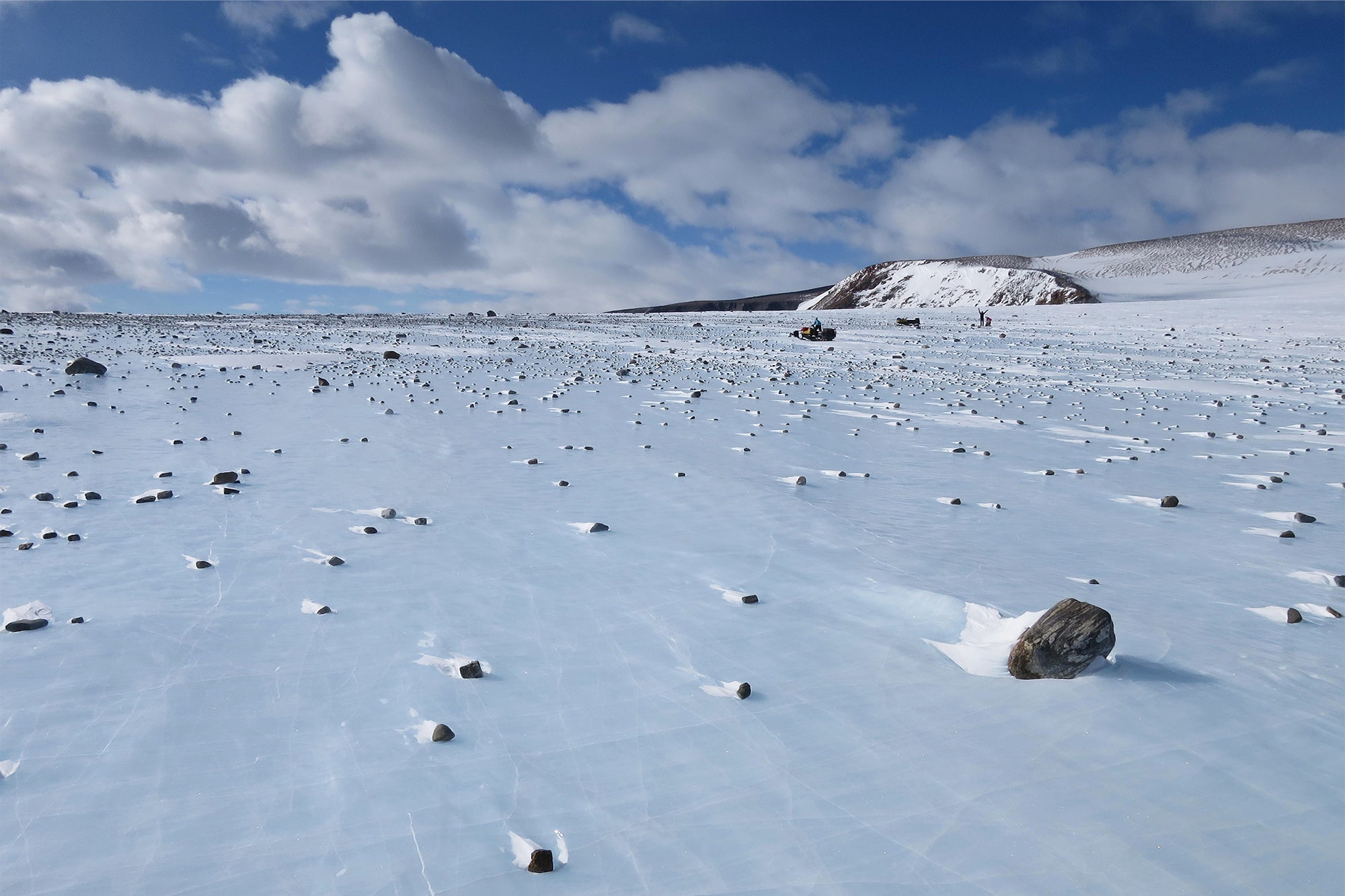 Daytime photograph of an ice field in Antarctica where dark colored meteorites strongly contrast visually against the bright blue-ish white ice surface