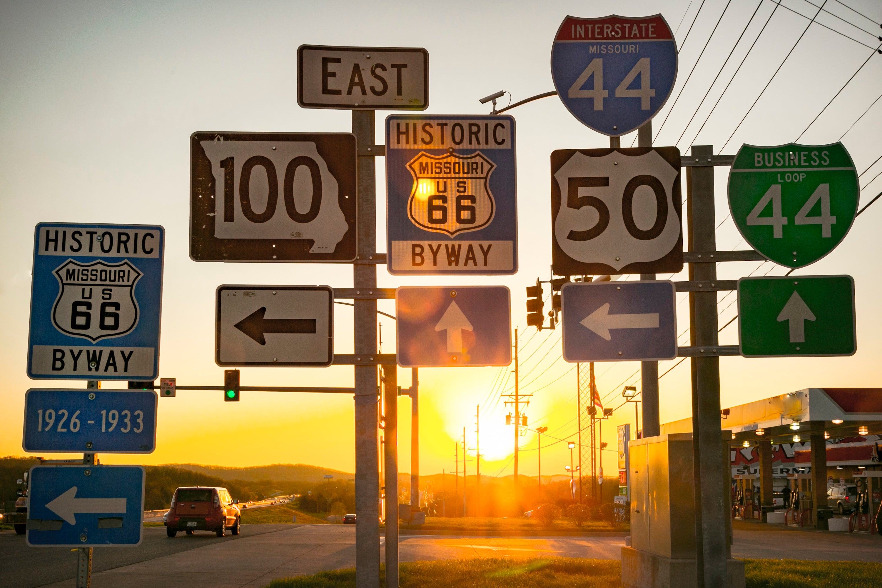 An array of about 12 road signs are in the foreground, with the open road leading toward sunset in the background.
