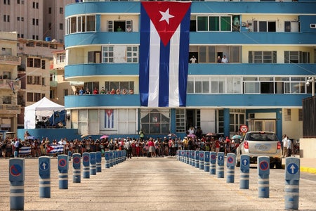 A crowd of people in front of a building watch a Cuban flag-rasing ceremony