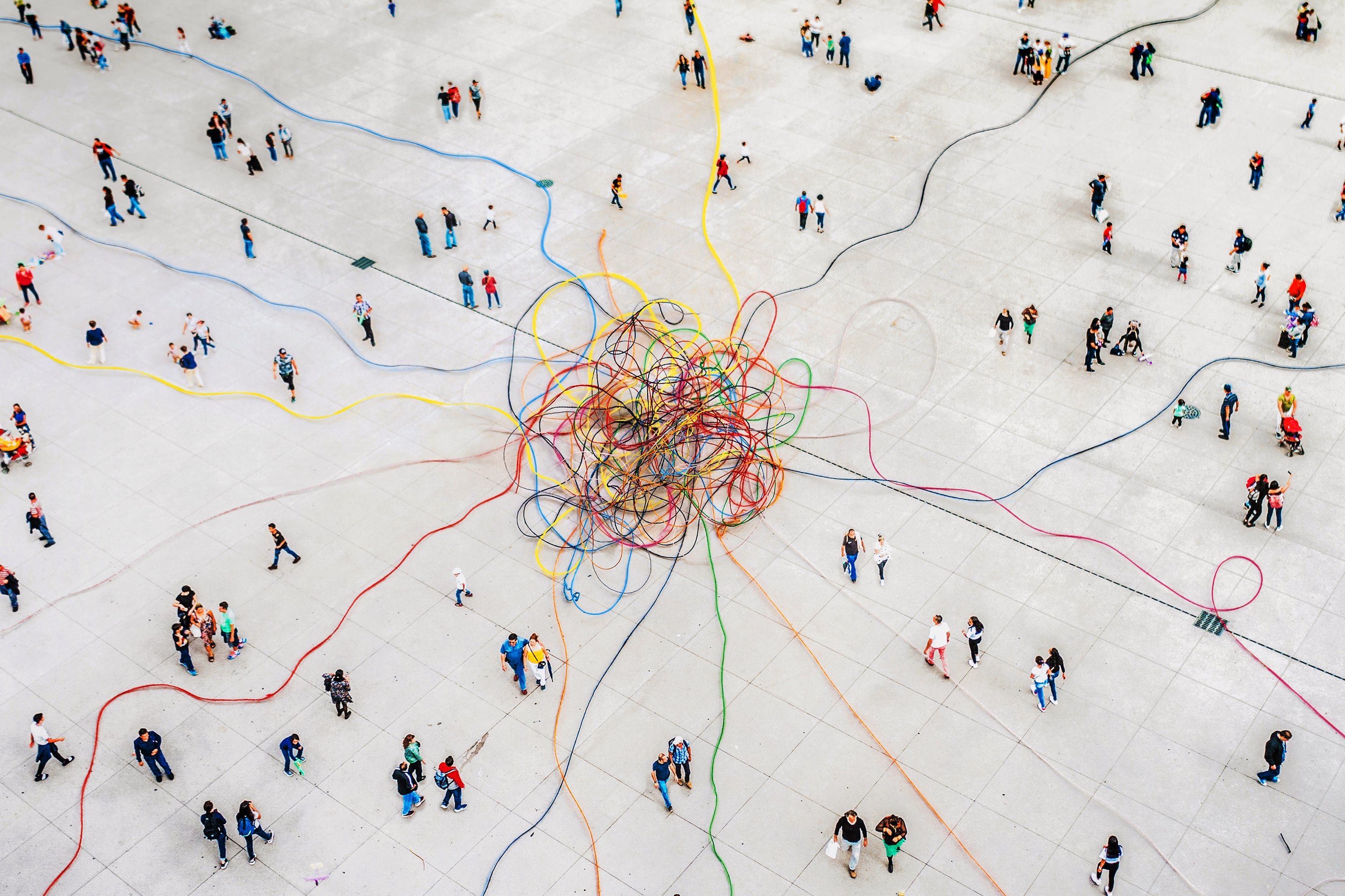 Pedestrian Traffic Turns to Chaos at a Critical Angle, Mathematicians Find Photo collage illustration, wide aerial view of crowd walking around a plaza with colorful threads of string leading from the outer edges towards the center where they are knotted together