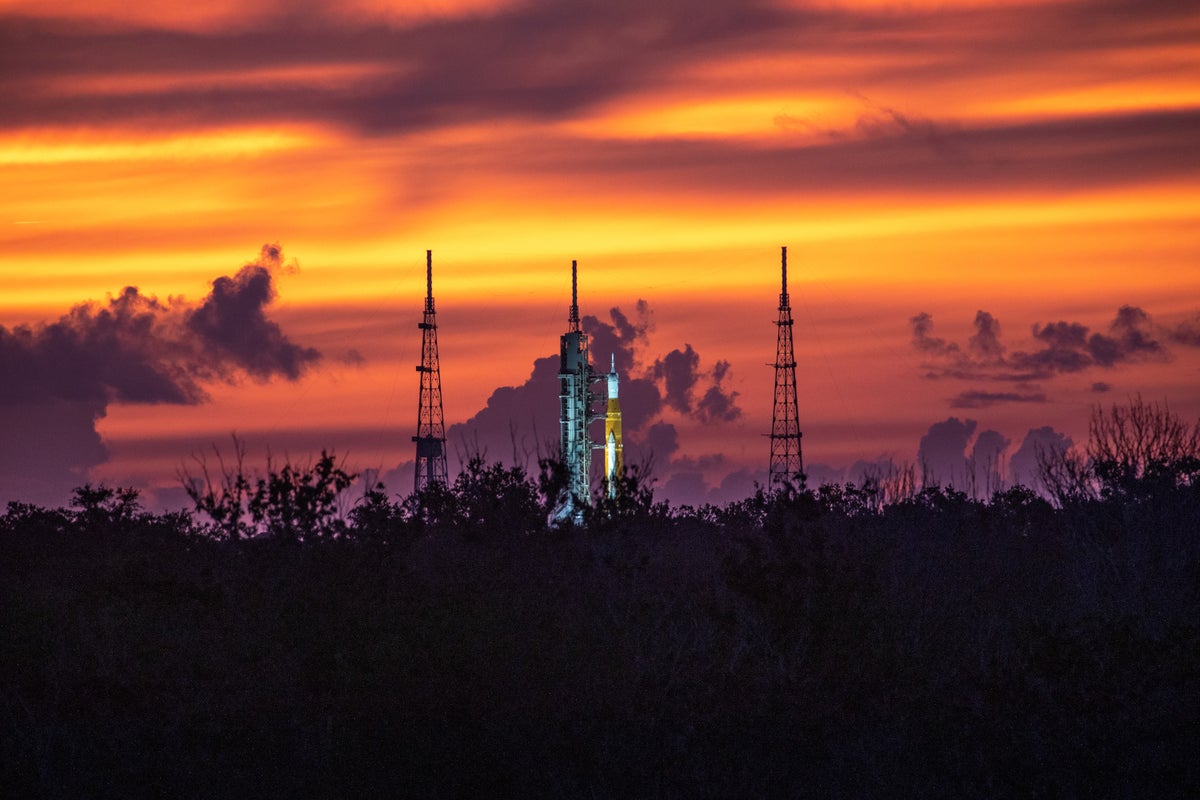 A space rocket stands bewtween two launch towers in front of a golden sunset.