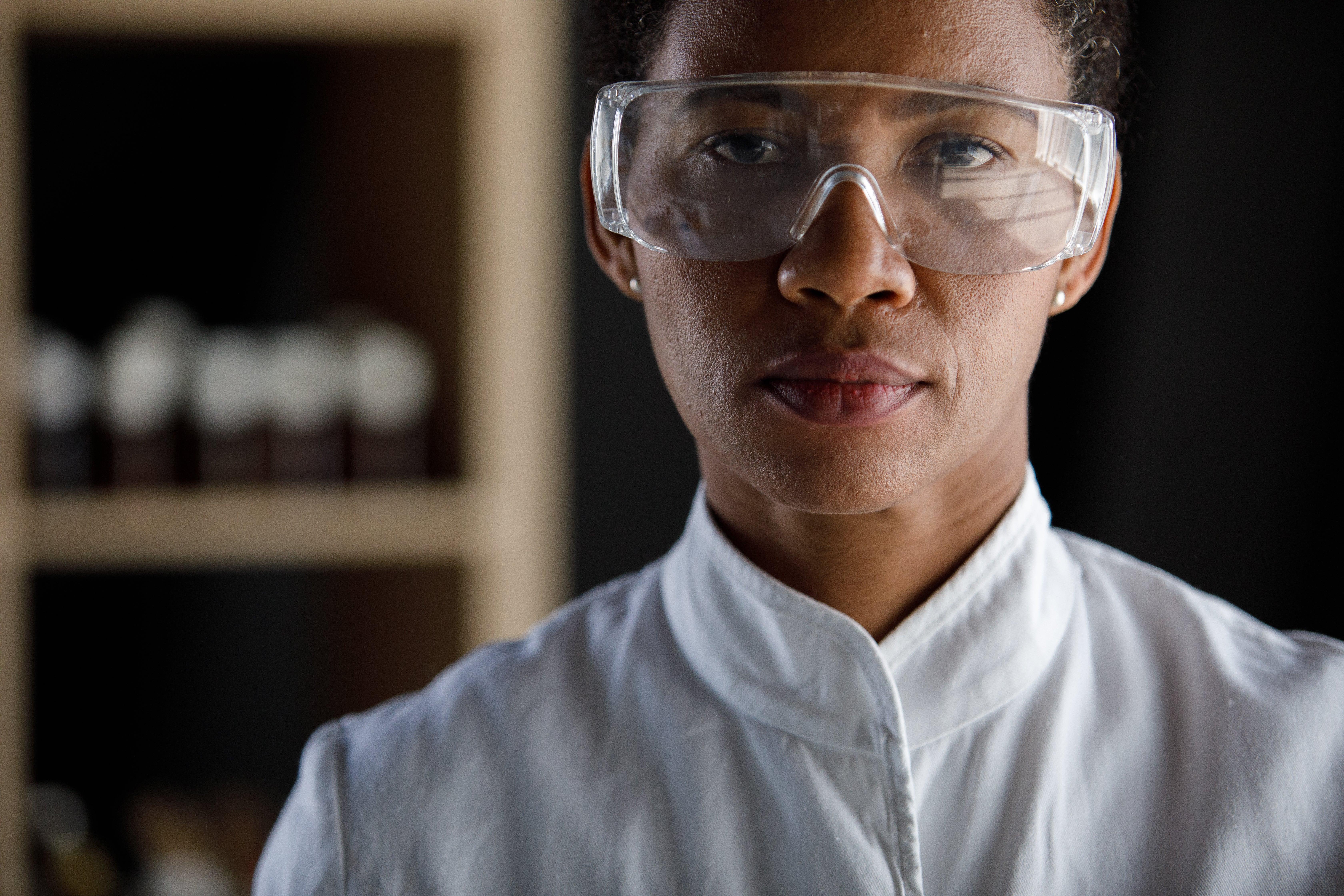 Serious woman of color scientist wearing protective eyewear in white coat.