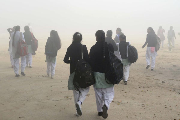 Backview of school girls in white pants and backpacks walking through smog on a sandy road.