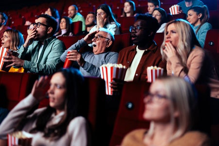 A diverse audience is in the movie theater watching a scary movie and gesturing in fear.