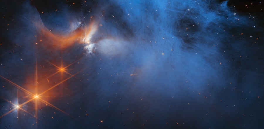 The cold, wispy cloud material (blue, center) in the central region of the Chamaeleon I dark molecular cloud is illuminated in the infrared by the glow of the young, outflowing protostar Ced 110 IRS 4 (orange, upper left). The light from numerous background stars, seen as orange dots behind the cloud, can be used to detect ices in the cloud, which absorb the starlight passing through them.