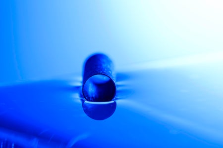 A short hollow metal tube floats horizontally on the surface of blue-tinted distilled water, with the open circular end facing the camera and reflected on the water below.