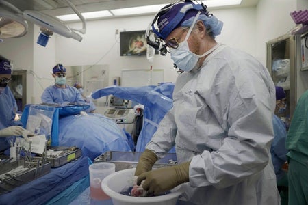 A man in surgical gear holds something in a bowl against a background of an operating room.