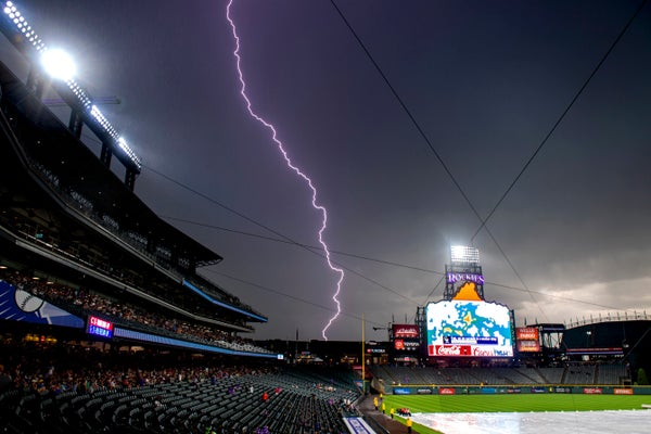 Lightning strikes (thin purple zig zag line) in purple sky of a baseball field between stadium seats to the left and green field to the right with display monitor in center