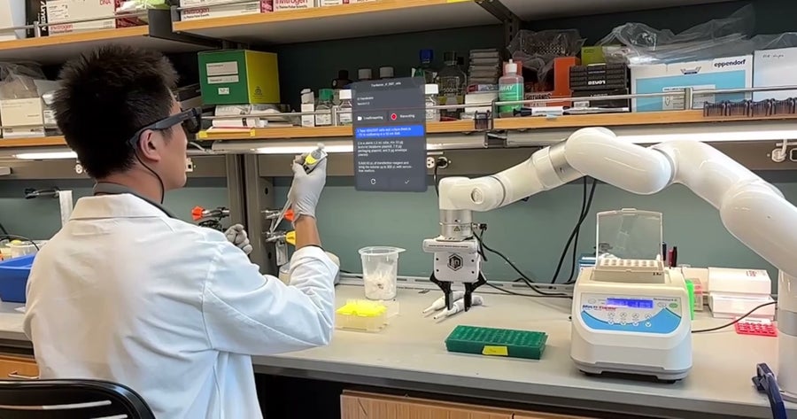 شخص يرتدي معطف المختبر، والقفازات، والنظارات الذكية يجلس على مقعد المختبر بينما يستخدم ماصة للتعامل مع أنبوب صغير. يتم وضع ذراع آلية على المقعد القريب. تحتوي الأرفف الموجودة بالأعلى على الصناديق ومستلزمات المختبرات. تعرض واجهة رقمية شفافة أمام الشخص خطوات البروتوكول ومؤشر التسجيل. يتم ترتيب معدات المختبرات، بما في ذلك الرف الأنبوبي وجهاز الطاولة، في جميع أنحاء مساحة العمل.