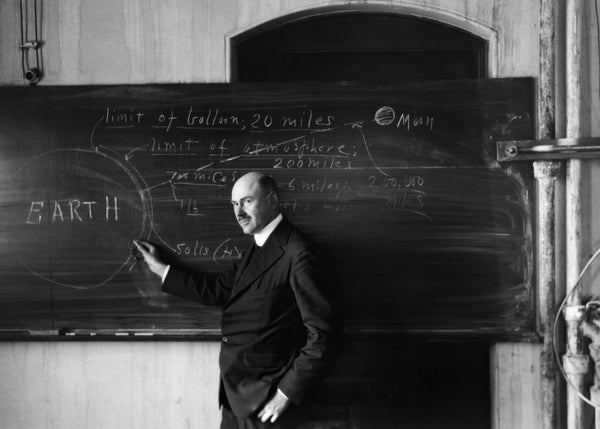A black-and-white photograph of a man standing in front of a blackboard holding a piece of chalk; a diagram of the Earth and moon is drawn on the blackboard.