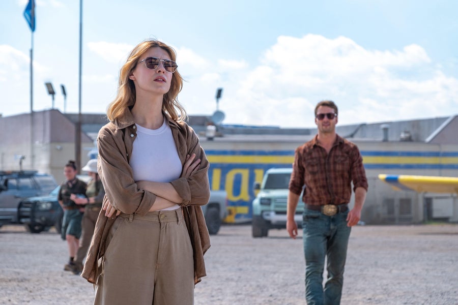 Daisy Edgar-Jones stands cross armed while Glen Powell walks towards her in the background in a film still from Twisters