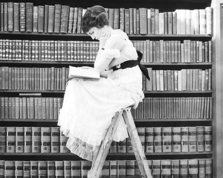 Young woman perched atop a ladder in library, reading.