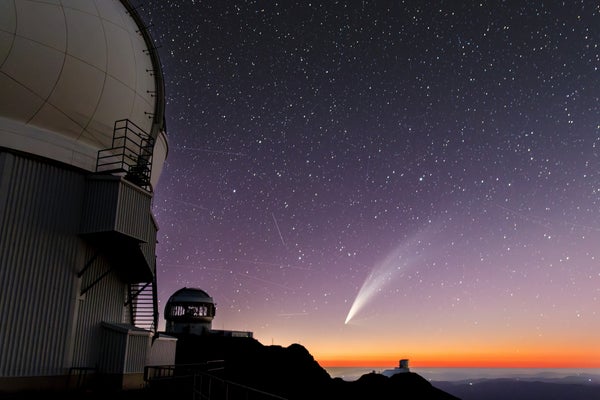 Kometa s propracovaným ohonem se ponoří do spodního středu snímku s astronomickými observatořemi v popředí.