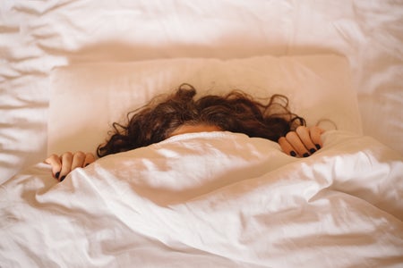 Woman hides under white blanket, only her long curly hair and dark manicured hands are visible