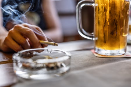 a hand holds a cigarette over an ashtray next to a beer on the table