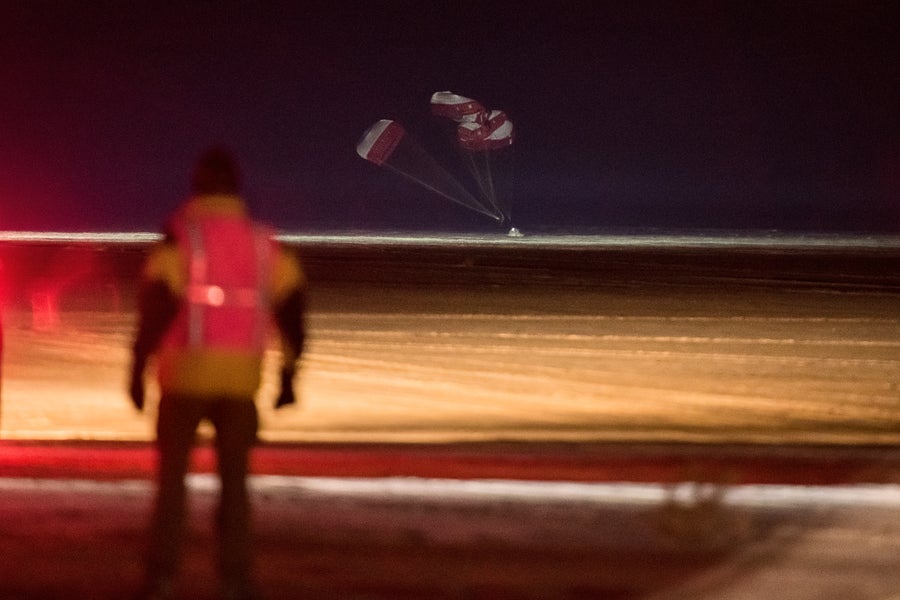 Des parachutes descendent au sol après l'atterrissage du Starliner de Boeing au Nouveau-Mexique lors d'un vol d'essai en 2019. Au premier plan, une personne se tient dos à la caméra et regarde le vaisseau spatial atterrir.