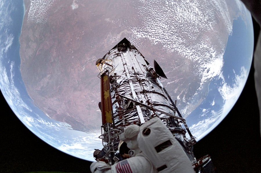 Photo, Shuttle STS-61 onboard view of the Hubble Space Telescope. Backdropped against planet Earth, the Hubble Space Telescope is shown sporting new and modified solar arrays stowed against its side. A spacewalking astronaut is visible in the foreground of the picture.