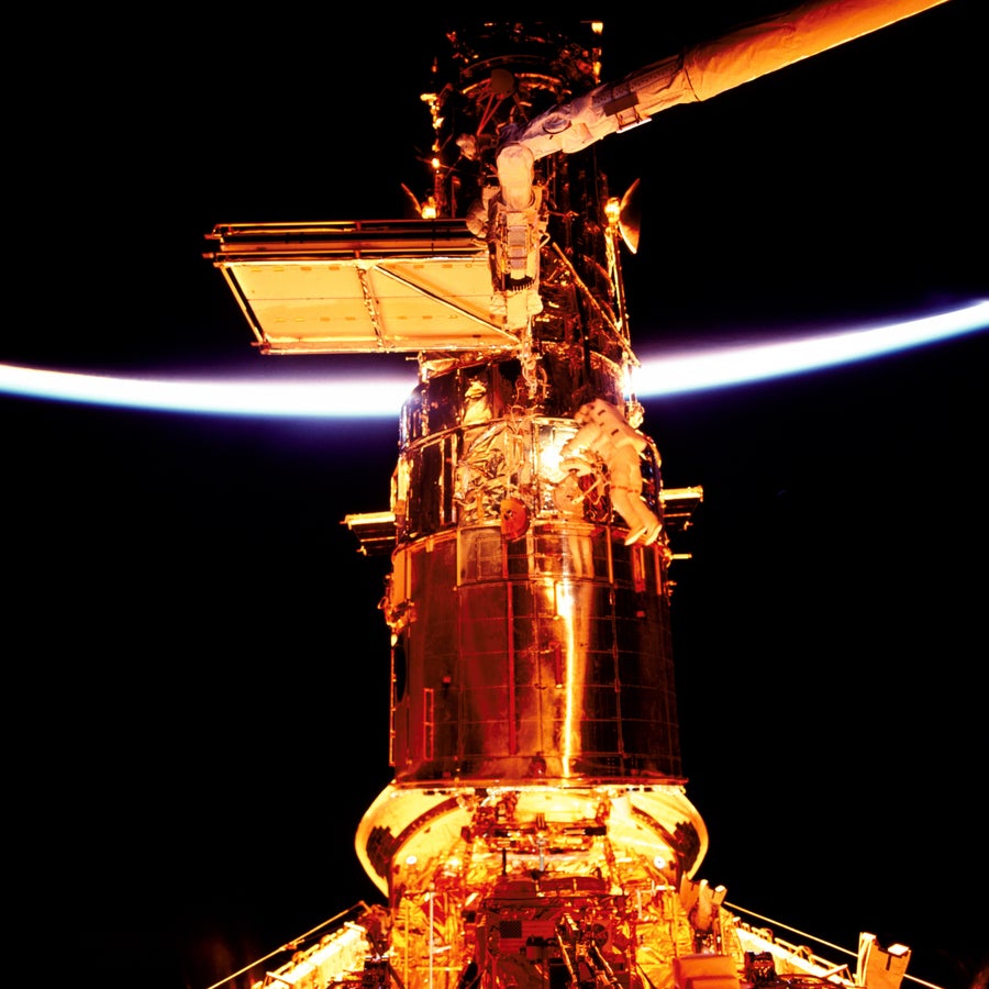 Two astronauts conducting a spacewalk alongside the Hubble Space Telescope. The sunlit edge of Earth is visible in the background.