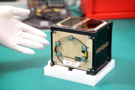 Wooden satellite box on a green tabletop with a white gloved hand presenting.