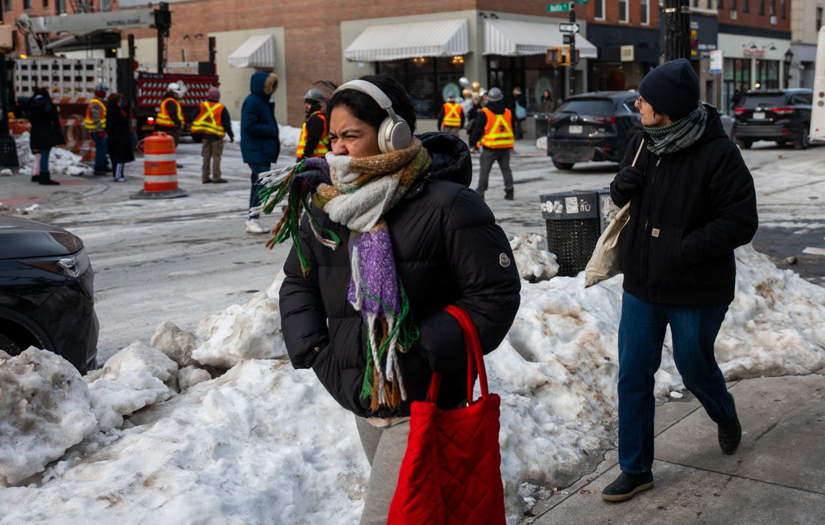 A person wearing a large black coat, colorful scarf and large headphones braces against a cold wind. They walk down a busy street in New York City where snow piles along the road.