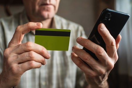 Elderly man win beige shirt at table holding up a credit card and phone.