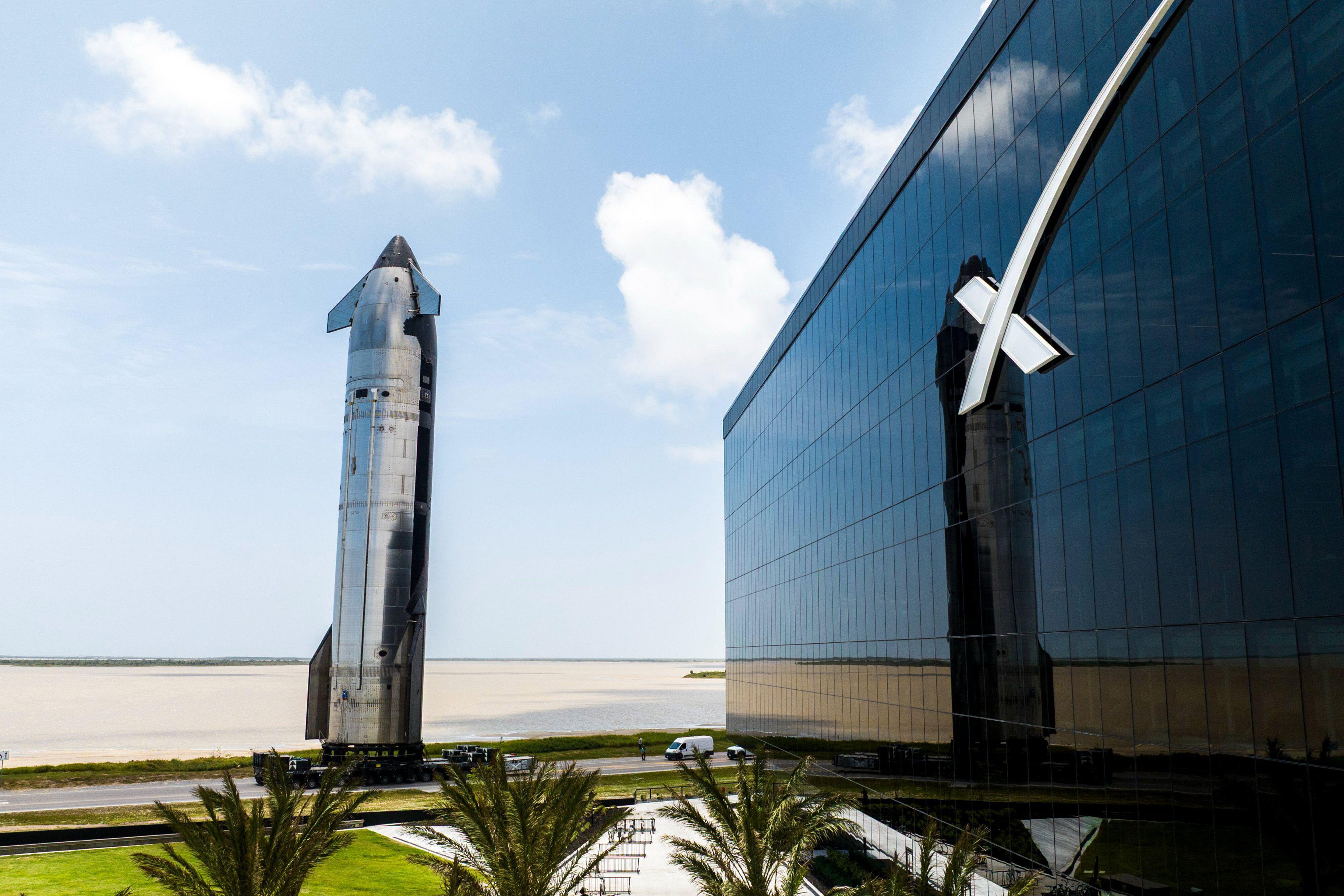 Massive silver rocket stands behind low industrial building at site