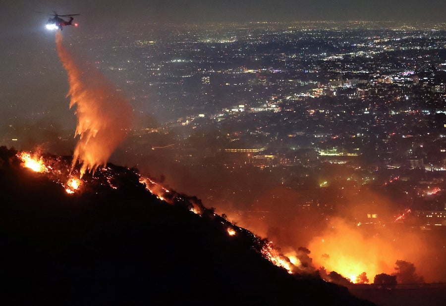A firefighting helicopter dropped water as the Sunset Fire burned in the Hollywood Hills with city lights visible behind the flames on a hill ridge as evacuations were ordered on January 8, 2025 in Los Angeles, California