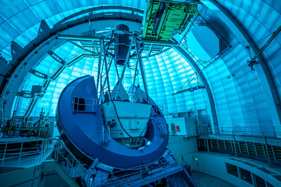 DESI’s instrument, a long black cylinder supported by the metal telescope framework, points at the domed ceiling. The dome interior is bathed in blue light.