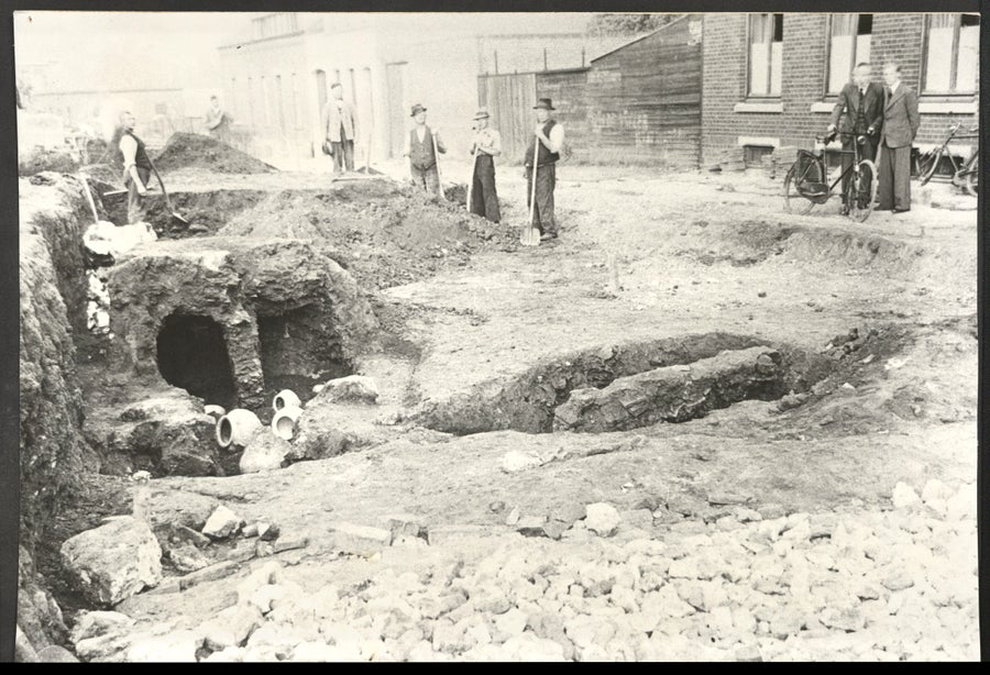 Black and white photo of pottery visible in a pit behind a pile of rubble. Bystanders look toward the pit