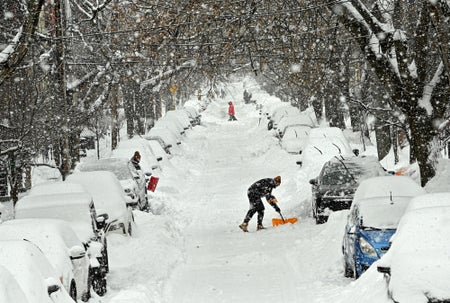 A snow-covered road with cars buried on either side going off into the distance. A person in the middle distance is shoveling out their car.
