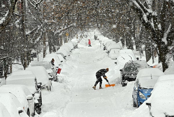 A snow covered road with cars buried on either side going far away. In the middle distance a person is taking his car out.