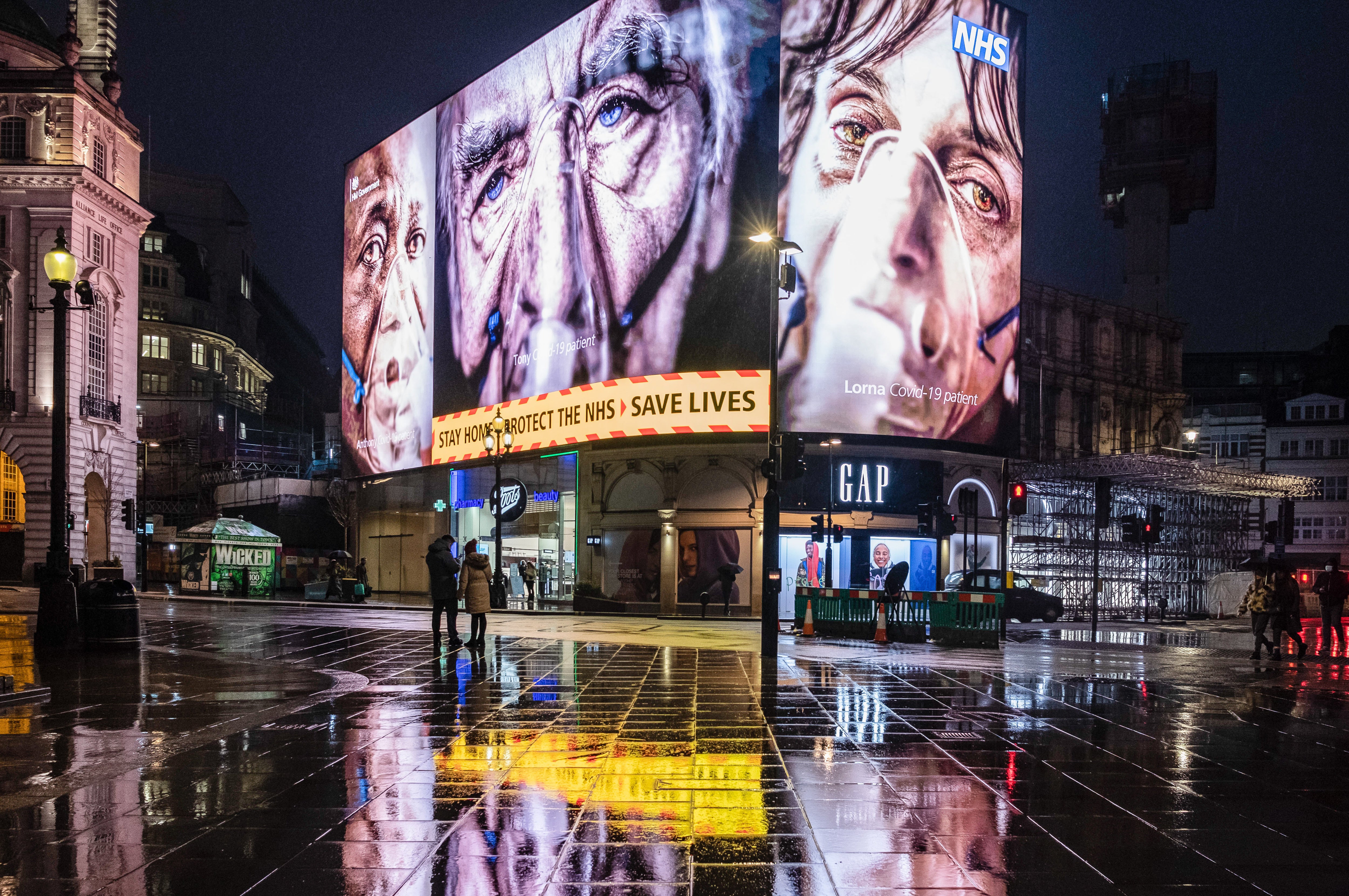 Silhouette of pedestrians walking past a COVID-19 electronic billboard showing three faces with oxygen masks, encouraging people to stay home, protect the NHS, save lives.