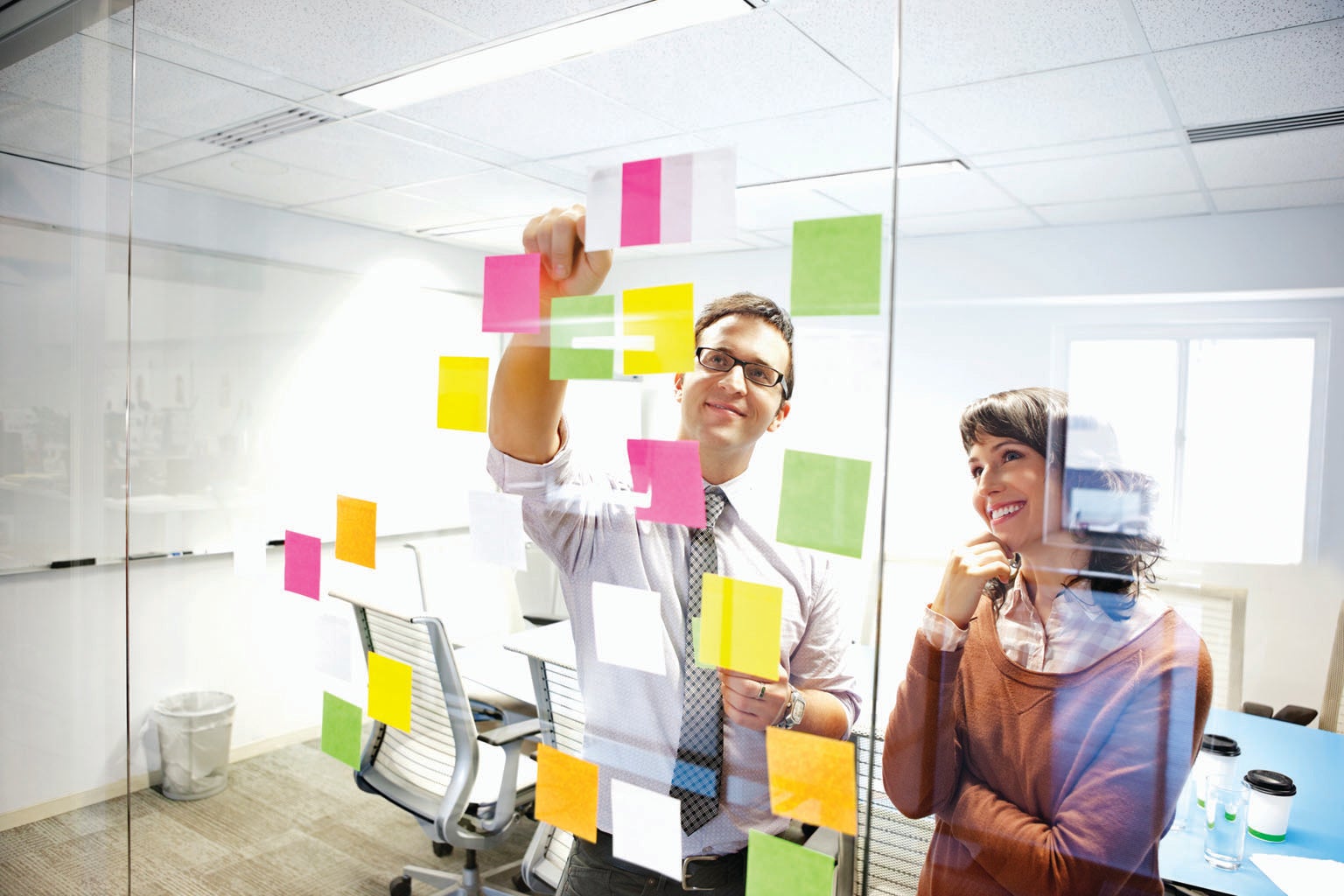 Man and woman smiling while posting notes on clear wall in office setting.