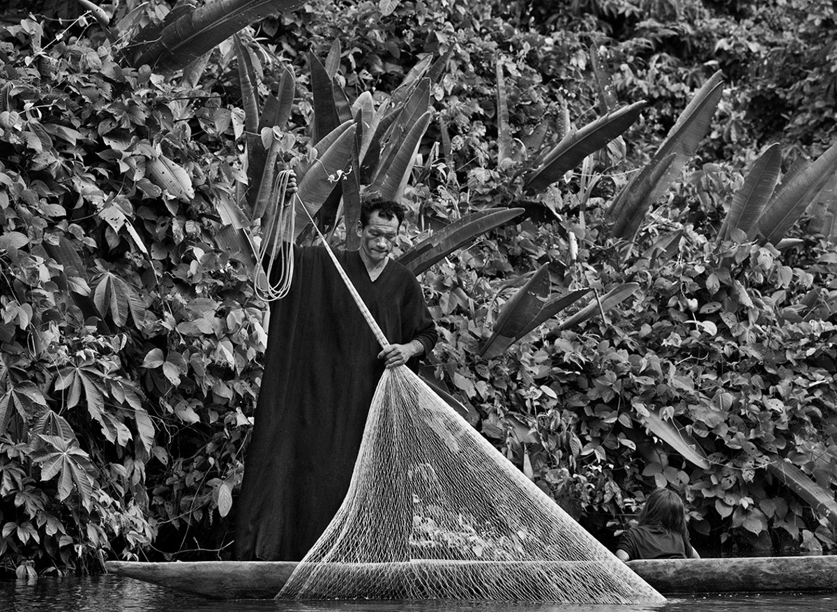 Man fishes by standing in a canoe and casting his net into the river.