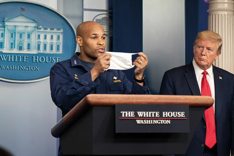 Former US surgeon general holds out a face mask stands next to Donald Trump during a White House briefing.