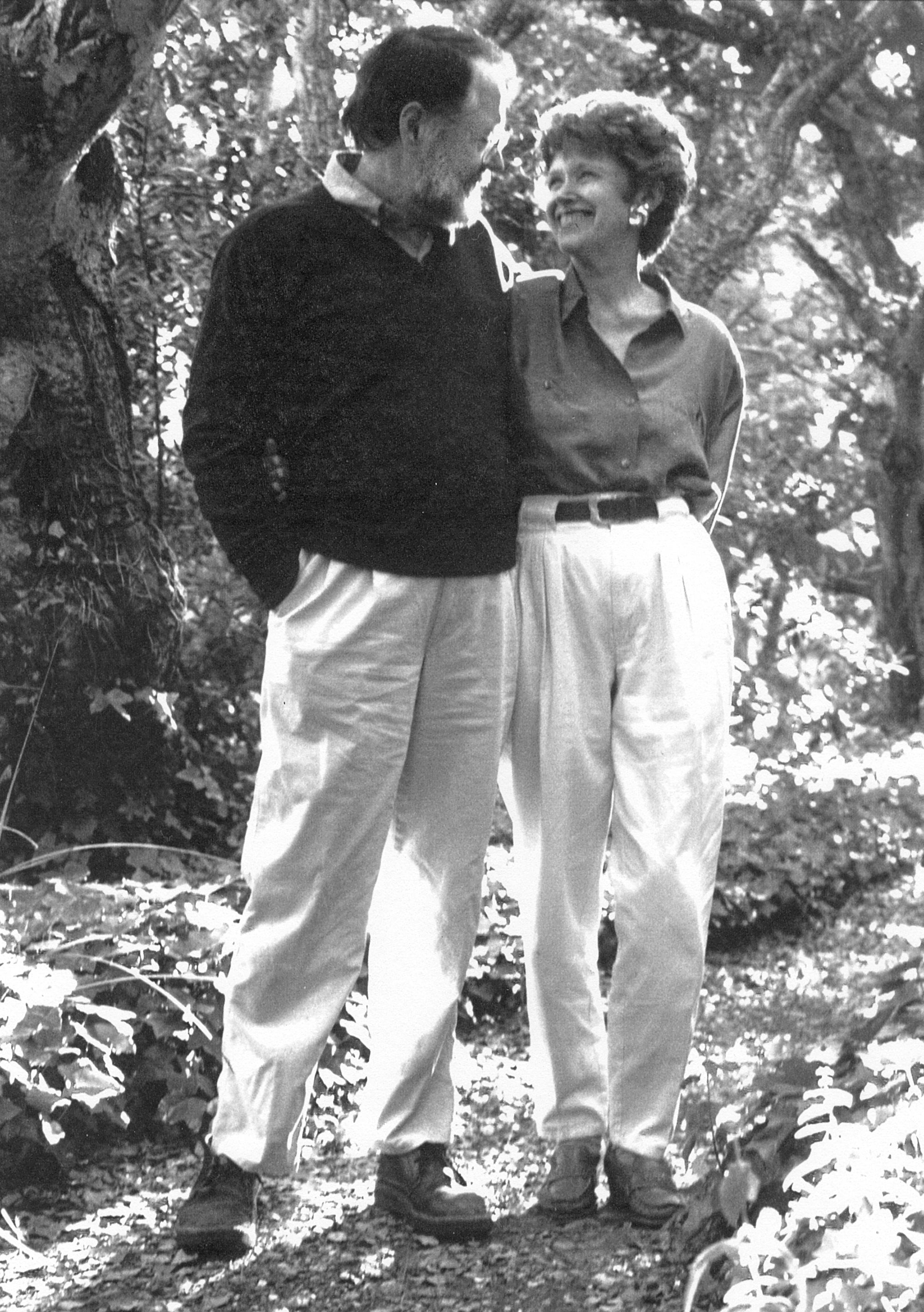 Black and white full-length portrait of man and woman in garden, looking at each other and smiling.