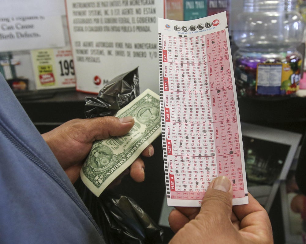 A customer in a liquor store waits in line to purchase a Powerball ticket while holding a dollar bill and a manually filled out Powerball form indicating the numbers they would like to play