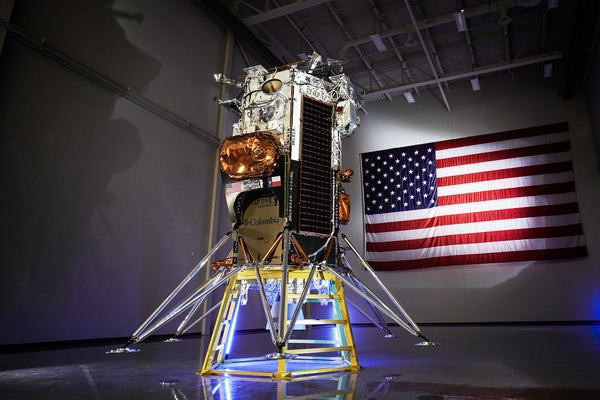 Intuitive Machines' Nova-C lunar lander on display in a cleanroom with a United States flag hanging on the wall behind the lander