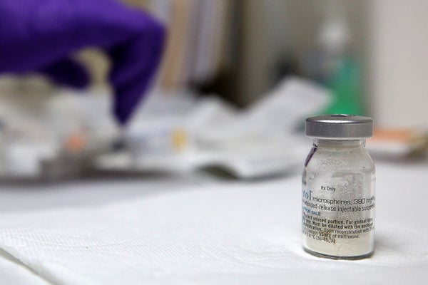 Close-up of a glass vial containing Vivitrol, a prescription drug, standing on a table. A nurse's gloved hand is seen in the background preparing injections