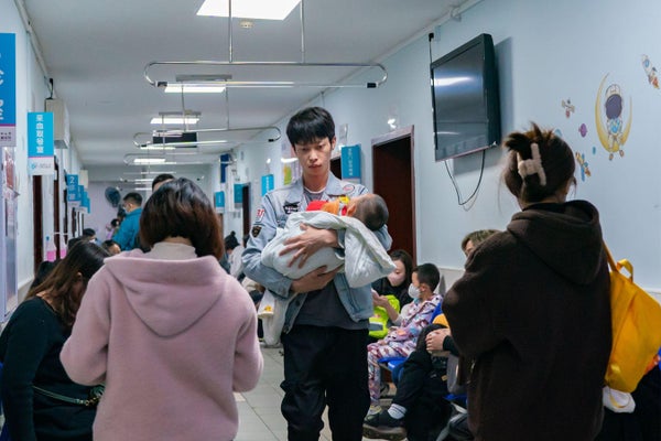Chinese man walking through a hospital waiting area with a baby in his arms - passing by seated waiting parents and children.