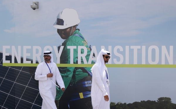 Men wearing thawbs walk past a billboard that reads ENERGY TRANSITION and shows a woman with hardhat and solar panels.